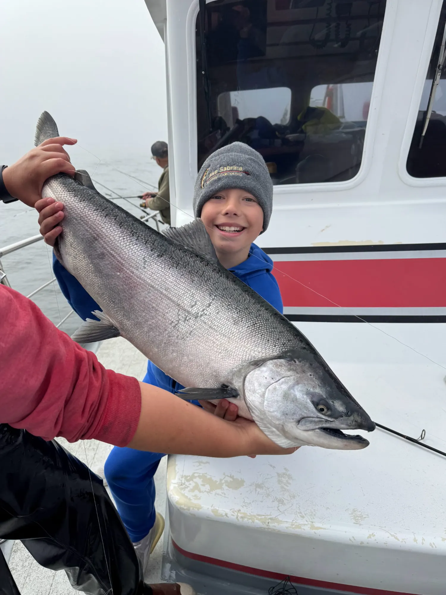 Chinook Salmon Gracie K 2005 Child smiling, holding a large Chinook salmon on a boat, wearing a grey beanie.