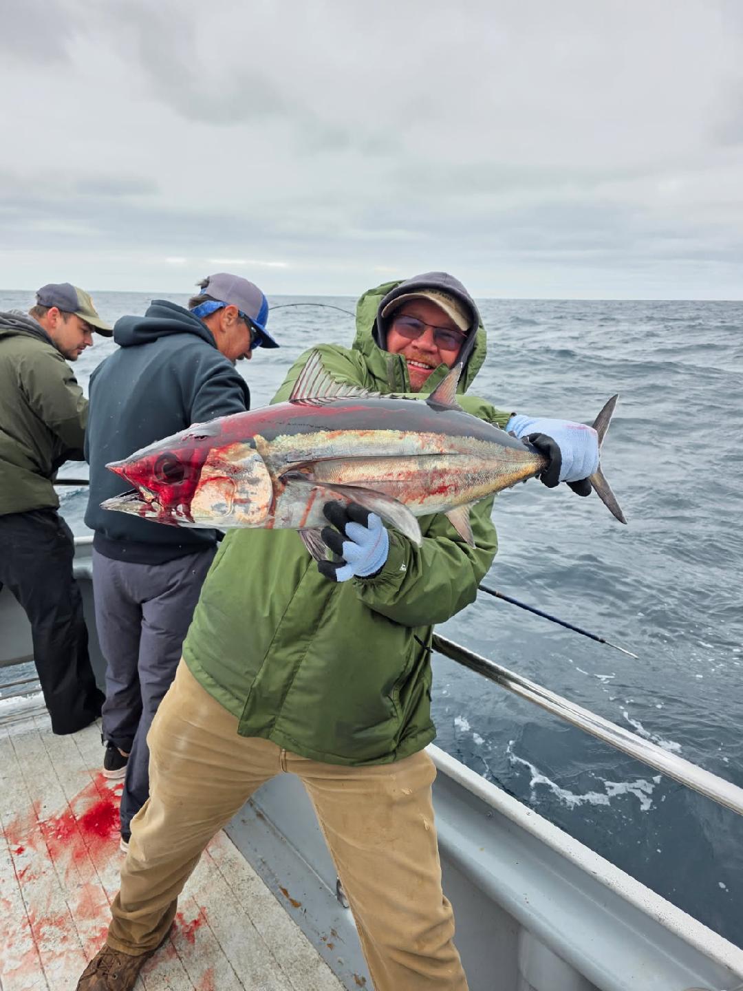 Man smiling holding up a freshly caught large albacore tuna with blood on it on a boat in the ocean